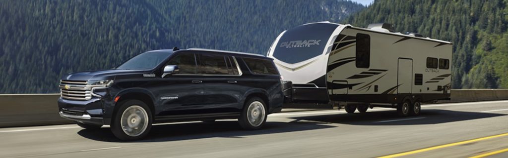 An all-new 2024 Black Chevy Suburban pulling a fifth wheel on a highway through the forest covered mountains of Sterling, VA.