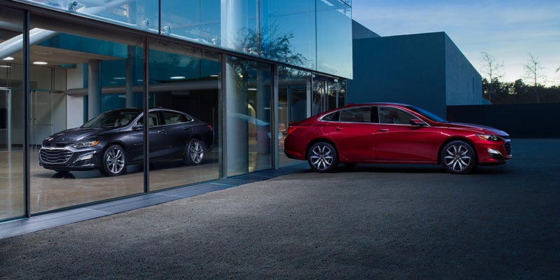 Two all-new 2024 Chevy Malibu's parked on opposite sides of a glass wall of a modern office building. The Malibu outside is Radiant Red Metallic and the Malibu inside is Lakeshore Blue Metallic. - Dealership in Sterling, VA
