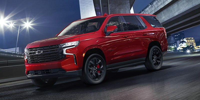 An all new 2024 Radiant Red Chevy Tahoe driving on the freeway below over-passes at night. - Ted Britt Chevrolet Sterling, VA