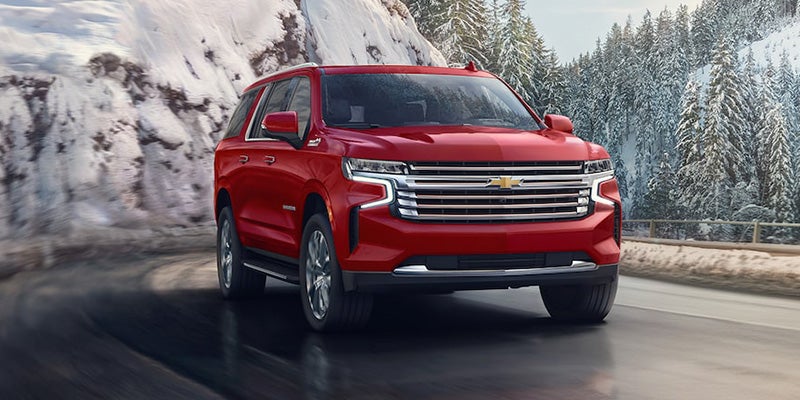 An all-new Red 2024 Chevy Suburban driving on a highway through some snow covered mountains. - Ted Britt Chevrolet Sterling, VA